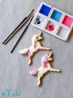 two decorated cookies sitting on top of a table next to paint and brushes with watercolors in the background