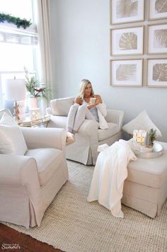 a woman sitting on a couch in a living room with white furniture and pictures above her