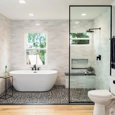 a white bath tub sitting next to a toilet under a window in a bathroom with wooden floors