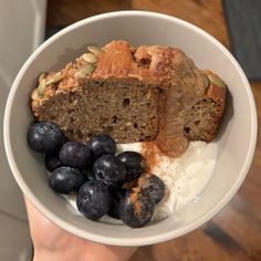 a person holding a bowl filled with blueberries and bread next to yogurt