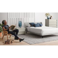 a woman sitting on a chair reading a book in front of a bed and dresser