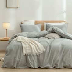 an unmade bed with grey linens in a white walled room next to a wooden headboard