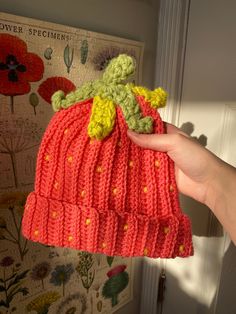 a hand holding a knitted strawberry hat in front of a wall with flowers on it