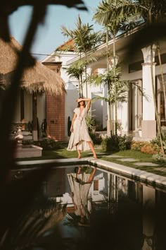 a woman standing in front of a pool with palm trees around her and a house behind her