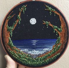 someone is holding up a small embroidery art piece with trees and the moon in the background