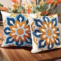 two pillows sitting on top of a wooden table with flowers in the vase behind them