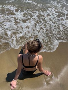 a woman sitting on top of a sandy beach next to the ocean with her feet in the water