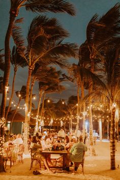 people sitting at tables under palm trees on the beach with lights strung from their branches