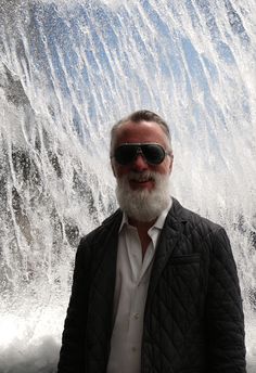 a man with a beard and sunglasses standing in front of a water fall that is spraying up into the air