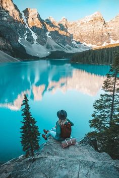 a person sitting on the edge of a cliff overlooking a lake with mountains in the background