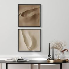 two framed pictures hang on the wall above a table with books and vases in front of it