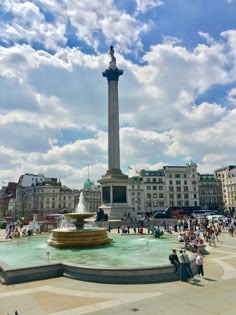 people are gathered around a fountain in the middle of a city square on a sunny day