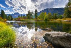 a river with rocks and grass in the foreground, surrounded by mountains on either side