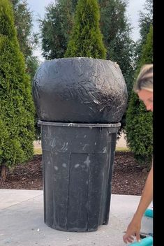 a woman standing next to a trash can on top of a cement sidewalk with trees in the background