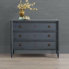 a black dresser with flowers on top in front of a gray wall and wooden floor