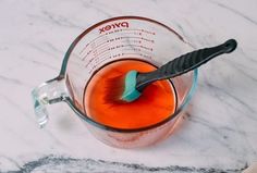 a toothbrush in a measuring cup on a marble counter top with red liquid inside