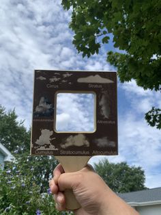 a hand holding up a square shaped object with clouds in the sky above it and trees below