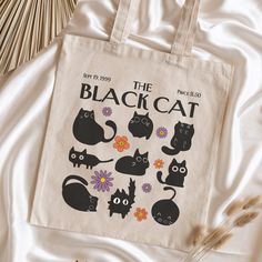 the black cat tote bag is sitting on a white sheet next to some dried flowers