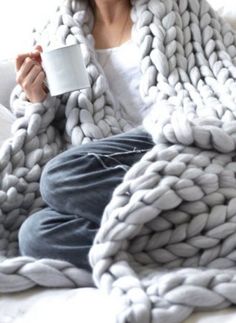 a woman sitting on a couch wrapped in a blanket holding a coffee cup and looking at the camera