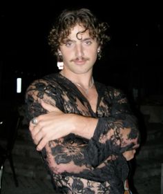 a man with curly hair wearing a black lace shirt and holding his arms crossed in front of him