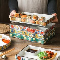 a person holding three trays filled with sushi and sauce on top of a wooden table