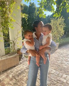 a woman holding two small children in her arms while standing on a brick walkway next to a tree