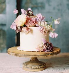 a white cake with pink and purple flowers sitting on top of a wooden stand in front of a brick wall