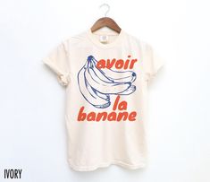 "Avoir la banane" is a French expression that means "to be in a good mood" or "to smile." It literally translates to "to have the banana". The expression is a colloquial way of saying that someone is happy or feeling great. We use professional quality DGT printing on all our apparel. Direct-to-garment, or DTG, is a high quality printing method that sprays ink directly onto the garment so there is no peeling or cracking. This fabulous graphic will be printed on a Comfort Colors Unisex T-Shirt. Pl Banana Shirt, French Quote, Quote T Shirt, Fruit Shirt, French Expressions, Good Mood, Printing Methods, Bananas, Comfort Colors