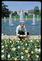 a man standing in front of a fountain surrounded by flowers and water with the white house in the background