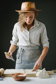 a woman in an apron and hat is cutting bread on a wooden board with a knife