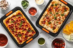 two pizzas on trays with toppings next to bowls of vegetables and condiments