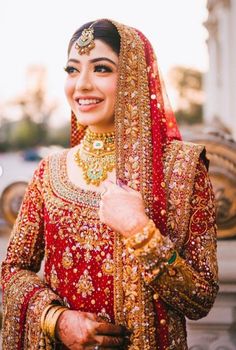 a woman in a red and gold bridal outfit smiles at the camera while standing next to a fountain