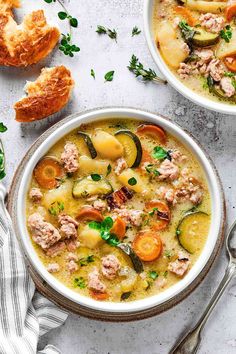 two bowls of soup with meat, vegetables and bread next to it on a white surface