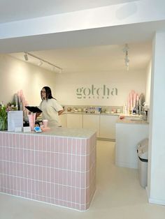 a woman standing behind a counter in a room with white walls and pink tiles on it