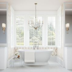 a white bath tub sitting in a bathroom next to two windows and a chandelier