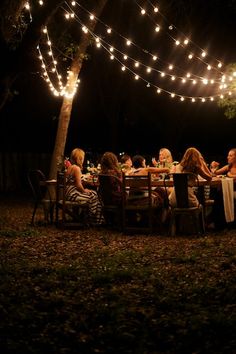 a group of people sitting around a dinner table at night with lights strung over them