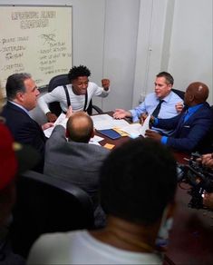 a group of men sitting around a table in front of a whiteboard with writing on it