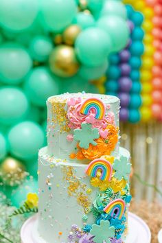 there is a white cake with rainbow decorations on the side and balloons in the background