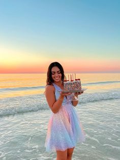 a woman standing in the ocean holding a cake with candles on it and smiling at the camera