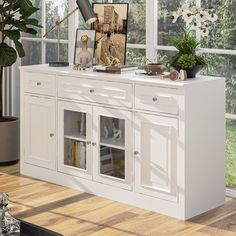 a white cabinet with glass doors in front of a window and potted plant on top