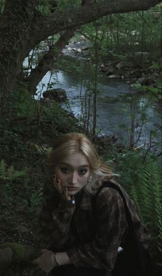 a woman sitting on the ground in front of a river with trees and bushes around her