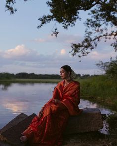 a woman in a red dress sitting on a rock by the water at sunset or dawn