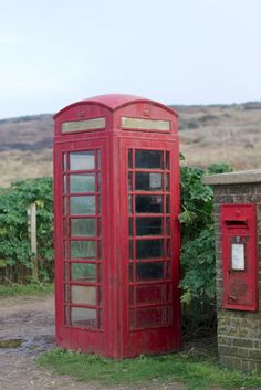 two red telephone booths sitting next to each other in the grass near a brick wall