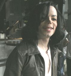 a man with long black hair wearing a leather jacket and white shirt smiling at the camera