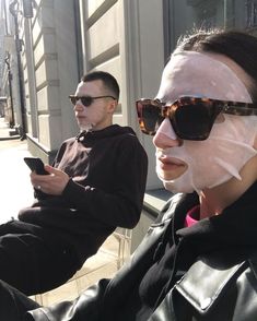 two people sitting on a bench with their faces covered in facial masks and looking at their cell phones