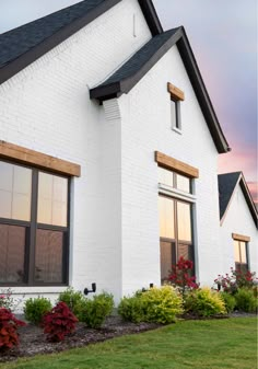 a white house with black shingles and red flowers in the front yard at sunset