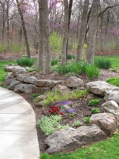 there is a rock wall in the middle of this yard with flowers and trees around it