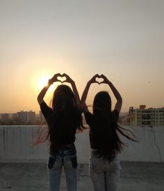 two girls making heart shapes with their hands in front of the sun setting behind them