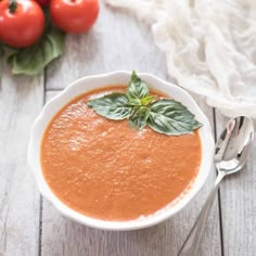 a bowl of tomato soup with basil leaves on top and tomatoes in the back ground