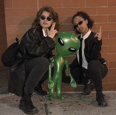 two women sitting next to each other near a brick wall with an alien doll in front of them